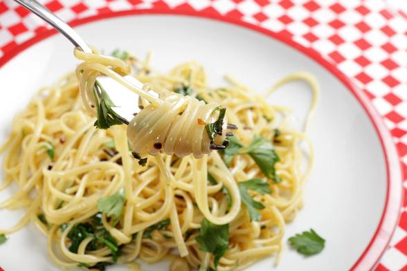 A white plate with a red border holds pasta with some green leafy bits. A fork twirls up some pasta. The background is a red and white checked tablecloth.
