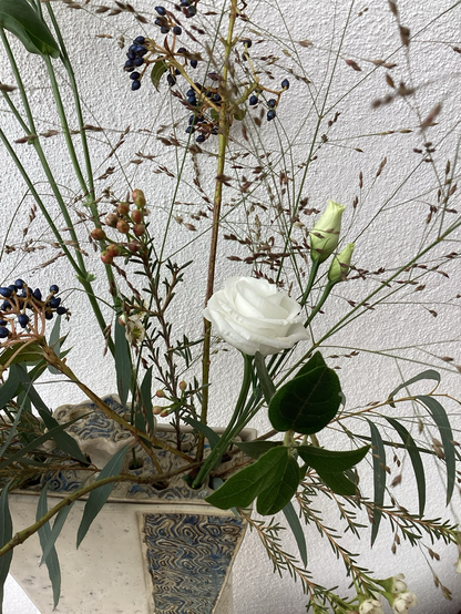 Links unten steht eine Tonvase mit einem durchlöcherten Deckel und einem Griff (beige mit blau/grauem Muster). Aus dem Deckel ragen sehr zarte Gräser und weiße Blüten.