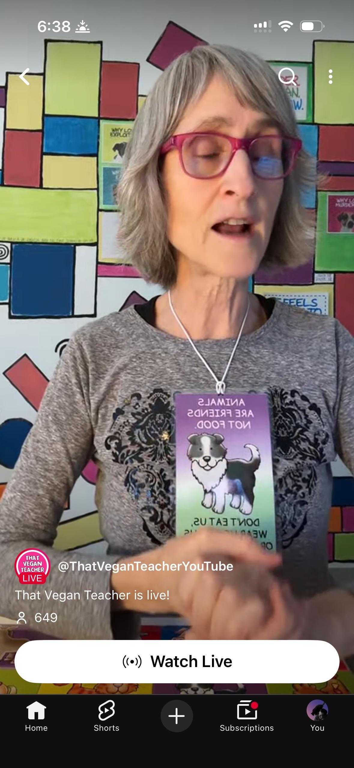 Older lady with short gray hair and red glasses. Wearing a gray shirt. Has a card around her neck with an illustration of a dog that says "Animals are not food"