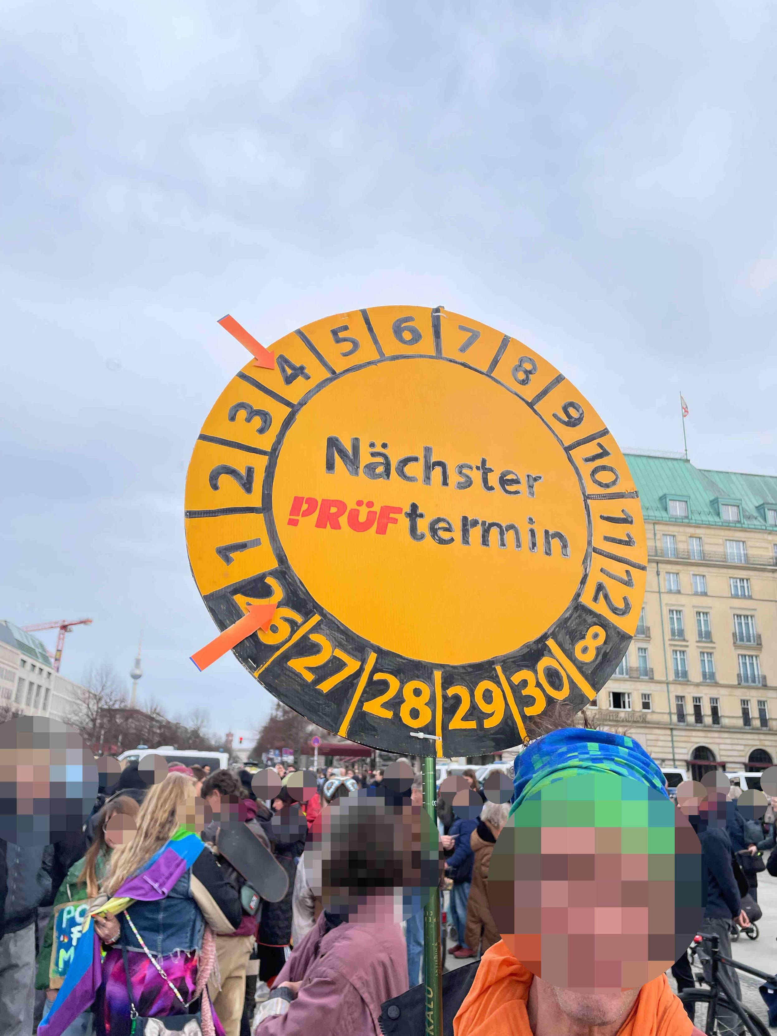 Demoschild wie Prüfplakette: Nächster Prüftermin 4/2026. Im Hintergrund verpixelte Demonstrierende.