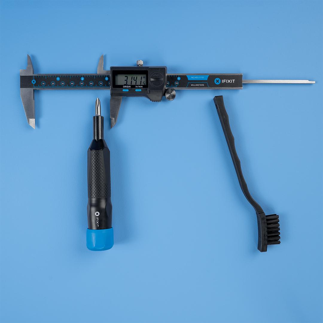 A digital caliper, iFixit precision screwdriver, and small cleaning brush are arranged on a blue background to resemble the Greek letter pi (ฯ), with the caliper forming the top bar and the two tools forming the legs.