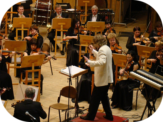 Un chef d'orchestre dirige un orchestre symphonique captivé, probablement lors d'une répétition ou d'un concert, avec une énergie palpable et une concentration silencieuse.
L'image présente une scène d'orchestre symphonique en pleine performance. Au centre, un homme aux cheveux bouclés et portant un costume blanc élégant, est visible de dos et de profil, semblant diriger l'orchestre avec des gestes animés. Il est le chef d'orchestre. Devant lui, une section de musiciens est positionnée, la plupart jouant des violons. Les femmes, principalement, sont assises avec leurs violons et leurs archets, concentrées sur leur jeu. Leurs tenues sont majoritairement sombres, peut-être des robes ou des tenues de soirée, contrastant avec le costume blanc du chef.
Derrière les violonistes, on aperçoit d'autres musiciens, dont certains jouent de cors d'harmonie, reconnaissables à leurs instruments cuivrés. Ces musiciens sont également assis et semblent porter des vestes sombres et des chemises claires, certains avec des nœuds papillon. À l'arrière-plan, on distingue des percussions, notamment un grand tambourin, et d'autres instruments moins clairement identifiables. Un clavier est également visible à droite de l'image, suggérant la présence d'un pianiste ou d'un claviériste.