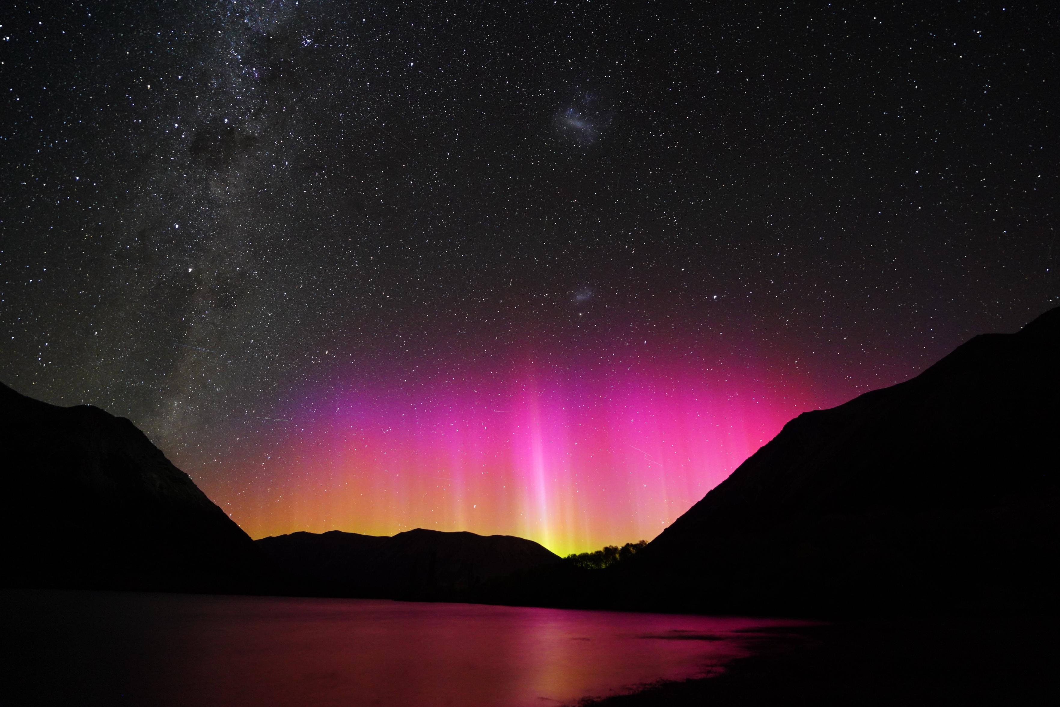 A nighttime view looking to the south, with the Milky Way stretching across the left portion of the photo, and the two Magellanic clouds visible as little puffs of cloudy stars, with blazingly bright pink and orange aurora pillars low on the horizon. The auroras are also visible reflected in a lake that's just barely visible in the starlight, with silhouetted mountains on each side of the image.