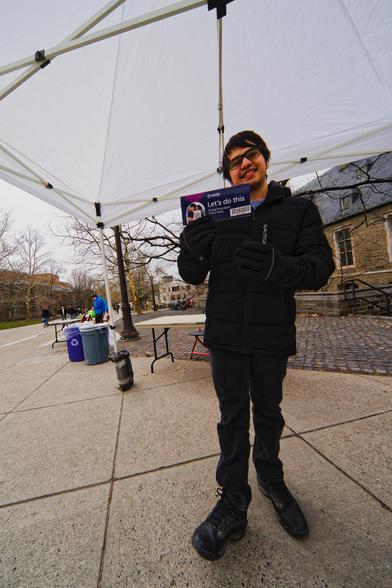 Man with dark hair and dark coat, gloves, pants and shoes holds a pamphlet that says "Let's do this. Saving lifes starts with a swab" as he smiles under a square white tent with more tables and people tabling and trees, bikes, trash cans and stuff in the background