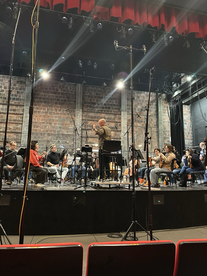 A still image showing a conductor standing on a podium surrounded by an orchestra of instrumentalists, a peppering of microphones throughout. The scene is backed by a large brick and cinderblock wall.