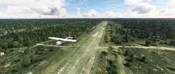 A small white and red single-engine propeller aircraft flies just above a narrow, straight concrete runway. The plane is slightly banked, suggesting it is taking off. The runway cuts through a dense, green forest that extends into the distance, with patches of open fields and clusters of trees visible on either side. The sky above is mostly clear, dotted with scattered clouds, and the landscape below appears flat and expansive, giving a sense of openness and natural surroundings.