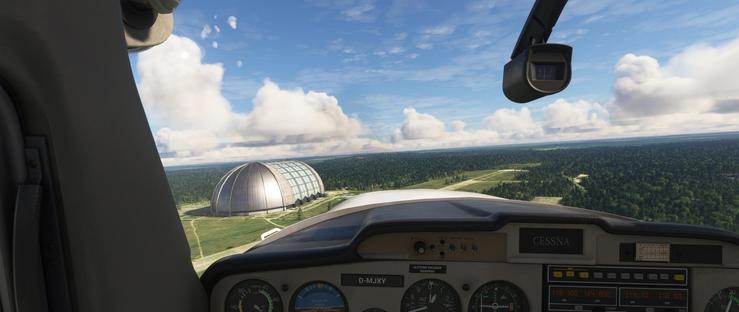 The cockpit of a Cessna aircraft shows the instrument panel with the registration D-MJXY visible. The airspeed indicator reads approximately 90 knots, and the altimeter, vertical speed indicator, heading indicator, and turn coordinator are clearly visible. Outside the windshield, a large, dome-shaped industrial structure sits amidst a forested landscape. The sky is partly cloudy, with patches of blue visible. The aircraft appears to be in level flight, passing near the structure, which stands out due to its size and metallic appearance. The surrounding area is dense with green forest, interspersed with clearings and fields. The horizon shows a mix of flat terrain and distant tree lines.