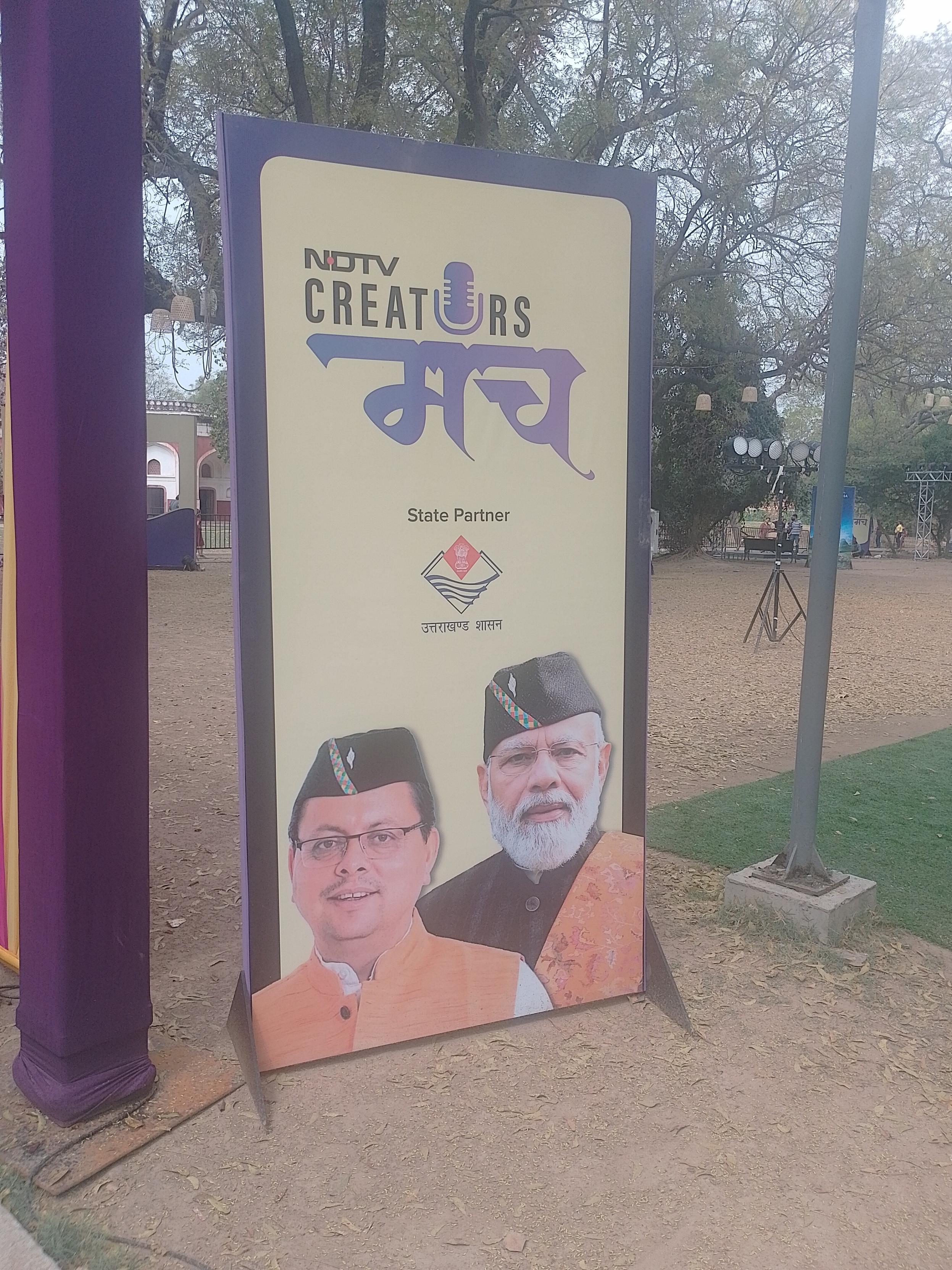 A large outdoor signboard with a beige background and purple border stands on a dirt ground in a park setting. The sign displays "NDTV CREATORS" with a microphone icon replacing the 'O' in "CREATORS", followed by the Devanagari word "मंच" in large purple script. Below this, "State Partner" appears above a logo and the Devanagari text "उत्तराखंड शासन" (Uttarakhand Government). The sign features two photographic portraits of men wearing traditional Uttarakhandi attire, including orange kurta and black caps with decorative bands. The person on the right is Indian Prime Minister Narendra Modi. The background includes trees, a purple pole, and park elements like a building and lighting fixtures.