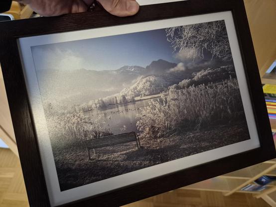 A framed photograph captures a serene and atmospheric winter lakeside scene, evoking a sense of peaceful solitude. The foreground features a wooden bench, positioned near the shore of a calm lake. The bench, lightly dusted with frost, invites quiet contemplation and offers a perfect spot to take in the stunning view.

The lake itself is partially frozen, its surface reflecting the soft, diffused light of the morning sun. Tall reeds and grasses, also covered in frost, line the shore, adding text…