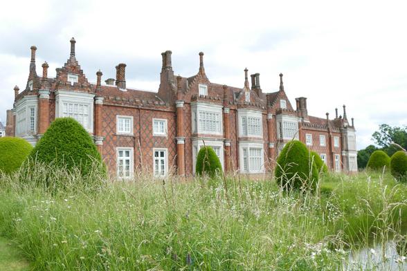 Akurat beschnittene kugelige Heckengehölze in einer wilden Wiese vor einem englischen Gutshaus.