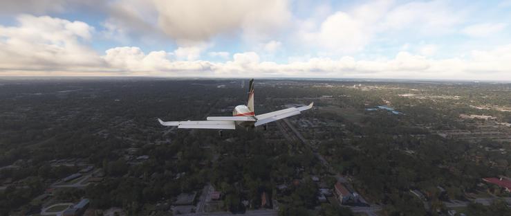 A small turboprop aircraft with a white fuselage and a dark vertical stabilizer featuring a red accent flies steadily above a suburban area. The plane’s landing gear is retracted, indicating it’s in flight, with its wings extending horizontally. Below, a dense pattern of residential neighborhoods unfolds, characterized by single-family homes with varied roof colors, winding streets, and patches of lush green trees. Larger wooded areas and open fields intersperse the housing developments, with a few water bodies reflecting light. In the distance, the landscape stretches toward a hazy horizon under a sky filled with scattered, fluffy white clouds. The lighting suggests a bright, clear day with sunlight filtering through the clouds, casting soft shadows over the terrain. The aircraft appears at a moderate altitude, offering a clear view of the detailed ground features beneath it, including streets, trees, and distant urban structures.