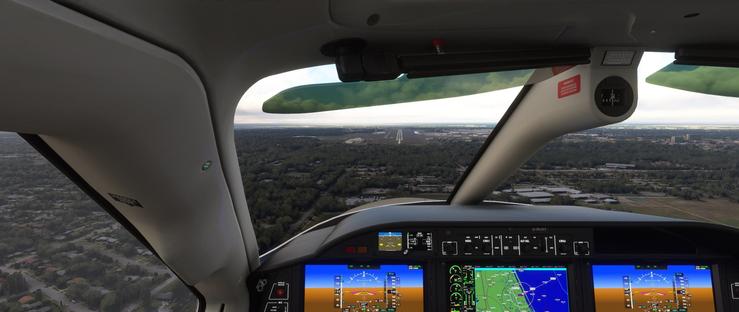 Airplane cockpit interior featuring three digital flight display screens. Left and right screens show altitude, airspeed, and heading data on blue and yellow backgrounds with color-coded meters, while the center screen displays a navigation map with green terrain, blue water, and a curved flight path. Below the displays, a control panel includes knobs and switches labeled ALT, HDG, VS, CRS, ALT SEL, CRS SEL, and BAND. Through the windshield, the aircraft is positioned above a runway, with surrounding landscape including dense green trees, suburban neighborhoods with houses and roads, open fields, and distant urban areas under a partly cloudy sky. A red warning label on the right side reads WARNING TURN ON, and a circular gauge near the top right of the windshield shows the number 24. The scene captures the cockpit’s layout and the aircraft’s approach toward the runway.