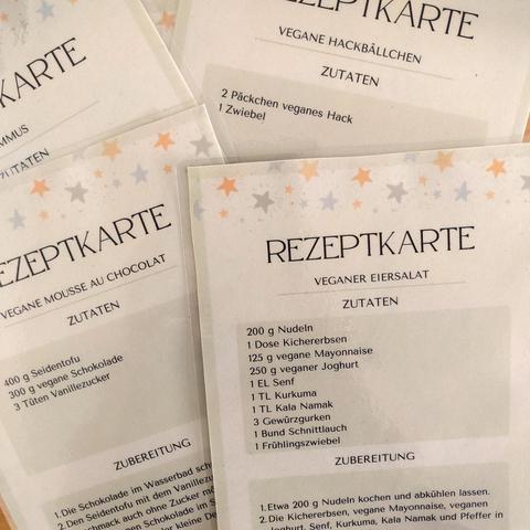 Mehrere Blätter Papier übereinander. Auf dem obersten steht ein Rezept für veganen Eiersalat. Ein anderes Blatt zeigt das Rezept für vegane Mousse au Chocolat. Die Rezeptkarten sind mit Sternchen dekoriert.