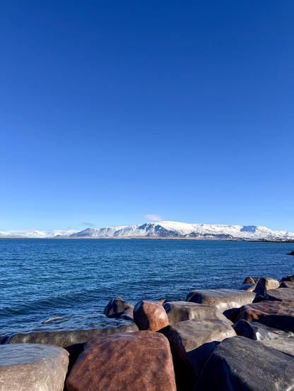 This image shows a rocky shoreline with smooth, rounded stones in the foreground and a vast expanse of deep blue water stretching out to the horizon. In the background, snow-capped mountains rise under a clear, bright blue sky.