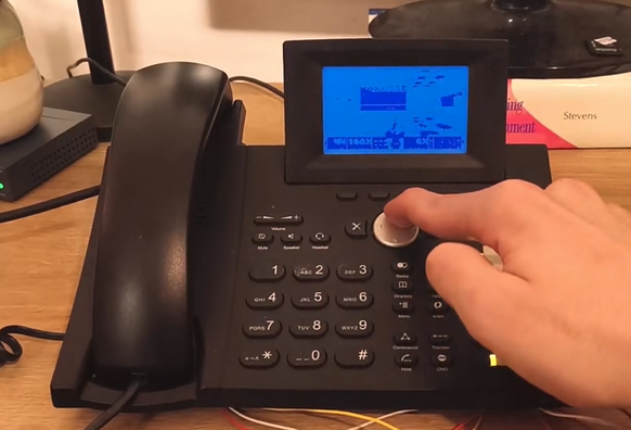 A low-quality image of the game Doom is visible on a small display with two shades of blue on a chunk black landline phone that has a person with a finger on the circular directional button.