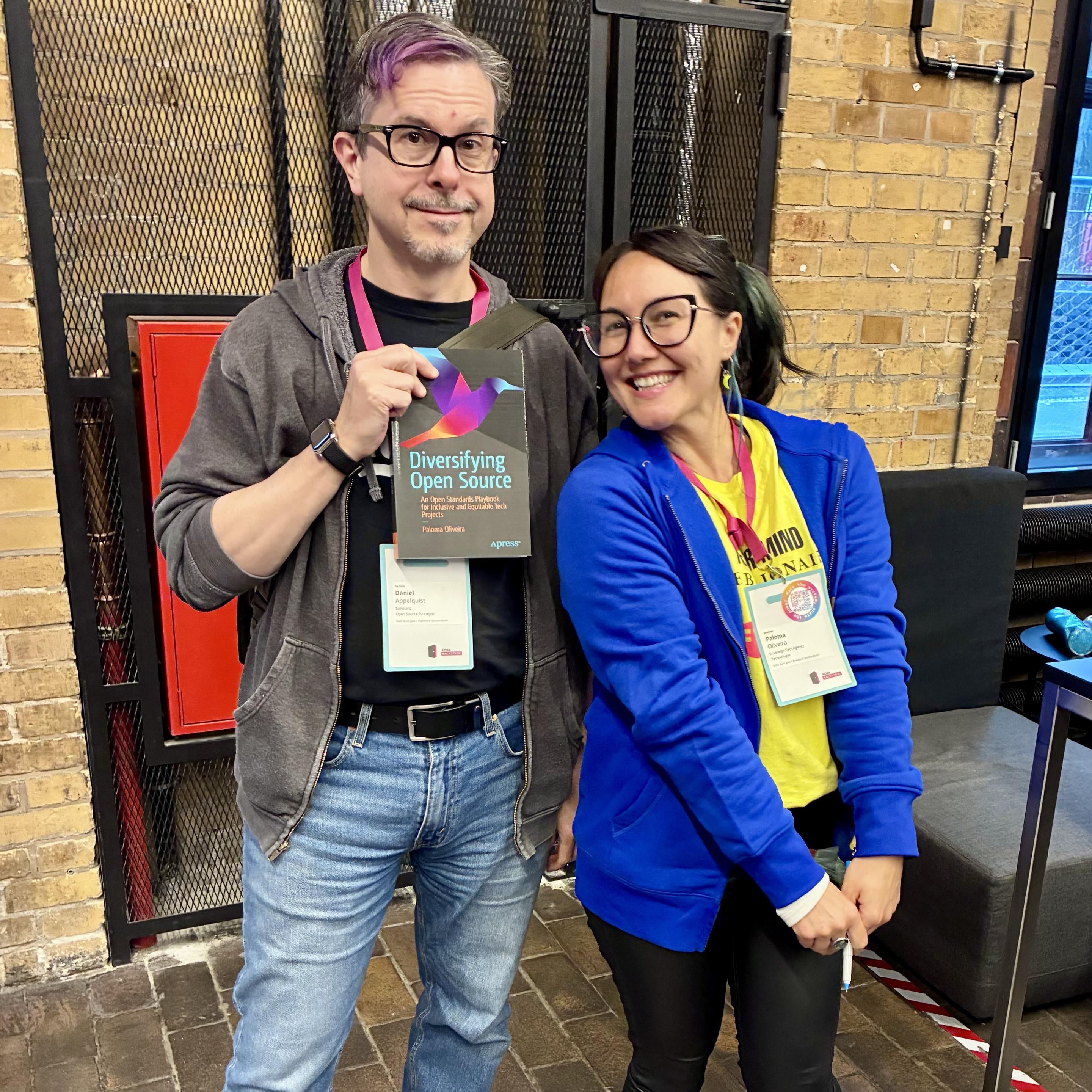 Paloma and me, smiling and posing together in an indoor setting, at the conference. I’m is holding the book titled "Diversifying Open Source." Both of us are wearing FOSS Backstage name tags, and there are brick walls and modern furniture visible in the background.