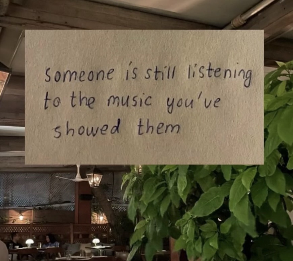 A photograph of a dimly lit, cozy restaurant interior with green plants. Overlaid on the image is a piece of paper with handwritten text that reads: "Someone is still listening to the music you've showed them."