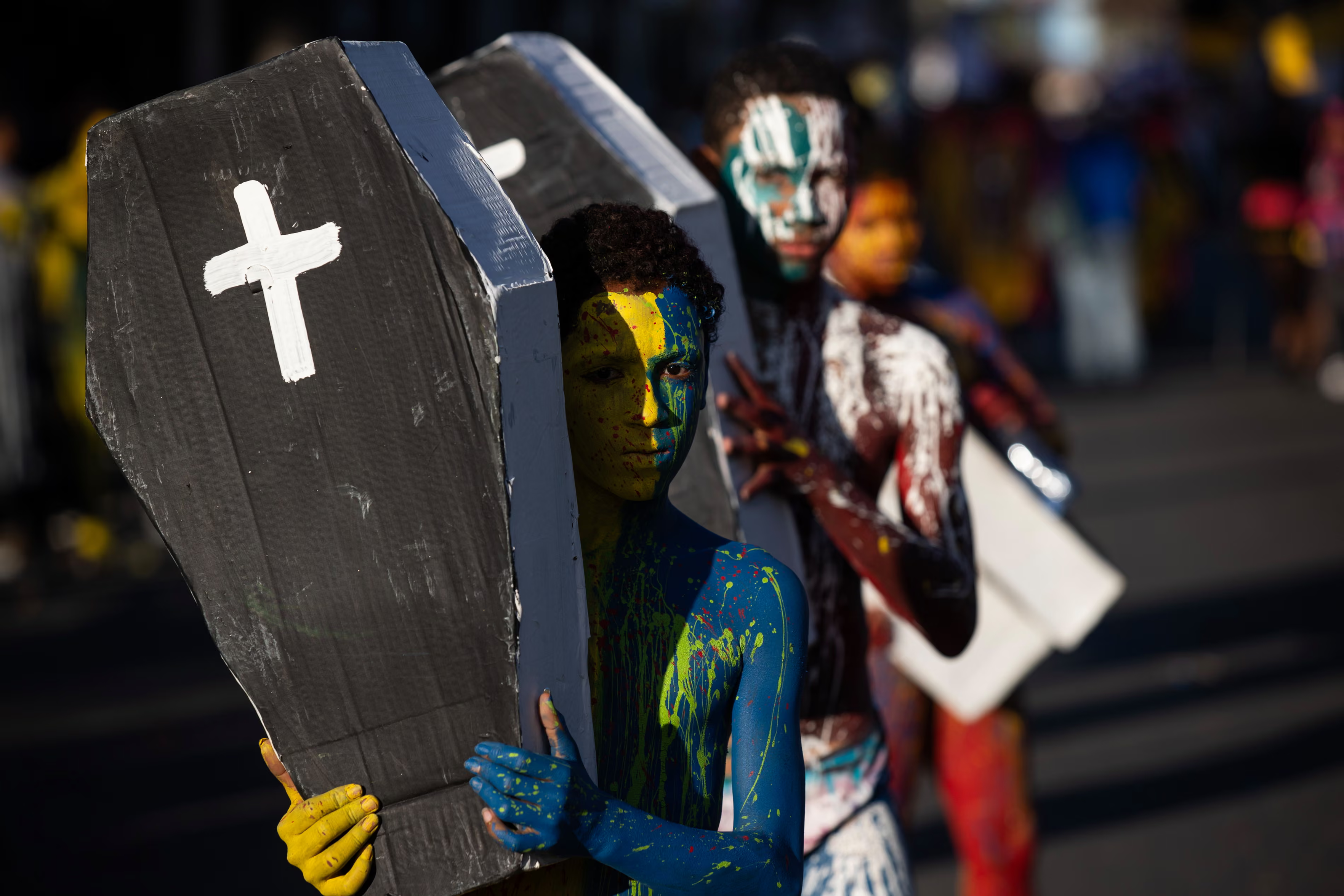 Children with pained bodies carry mock coffins with white crosses.