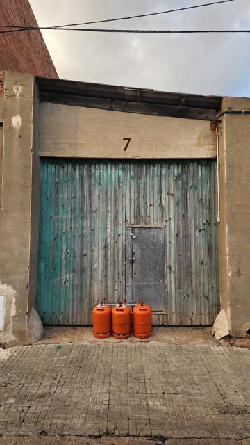 A large wooden gate, once painted gree, with a small metal door embedded. There are three small, dark orange, gas cylinders stamina in front of the gate. The house number seven is on the arch framing the gate.