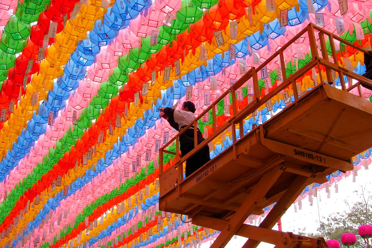 a person on the platform of a crane, reaching upwards to adjust rows and rows of colorful lanterns lined up according to colors