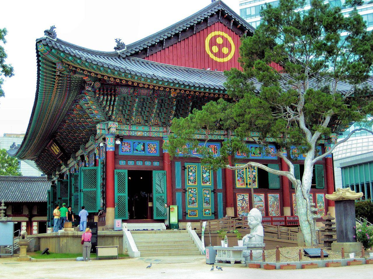 a building at the temple complex, it has blue-green roof and walls, some of the walls red, very colorful, seen from the front in a three quarter view, a tree and some sculptures in front of the building, some people walking around outside of it