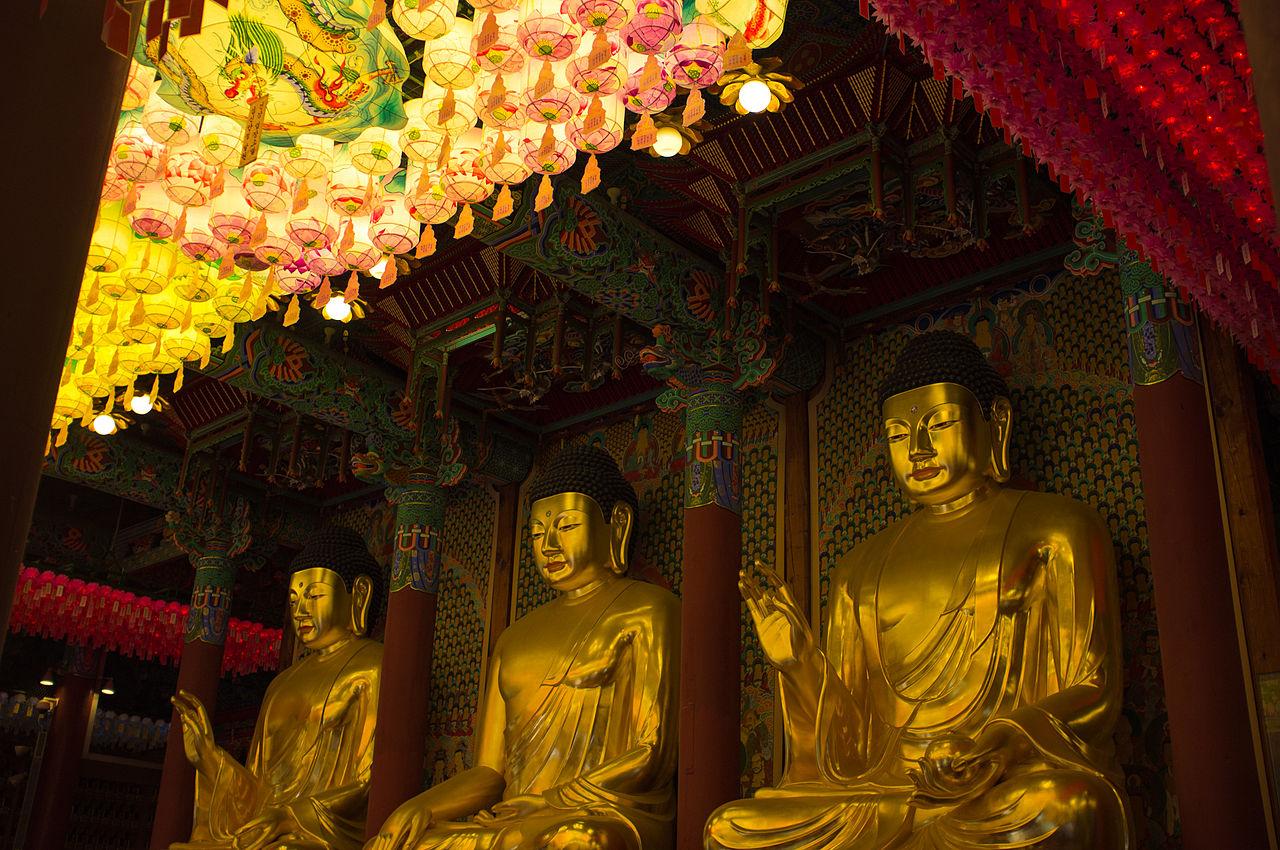 three golden buddhas sitting on the altar, glowing lanterns in front of them, two of the buddhas on the left and right are raising their right hands up, the center one has his resting on his knees palms down