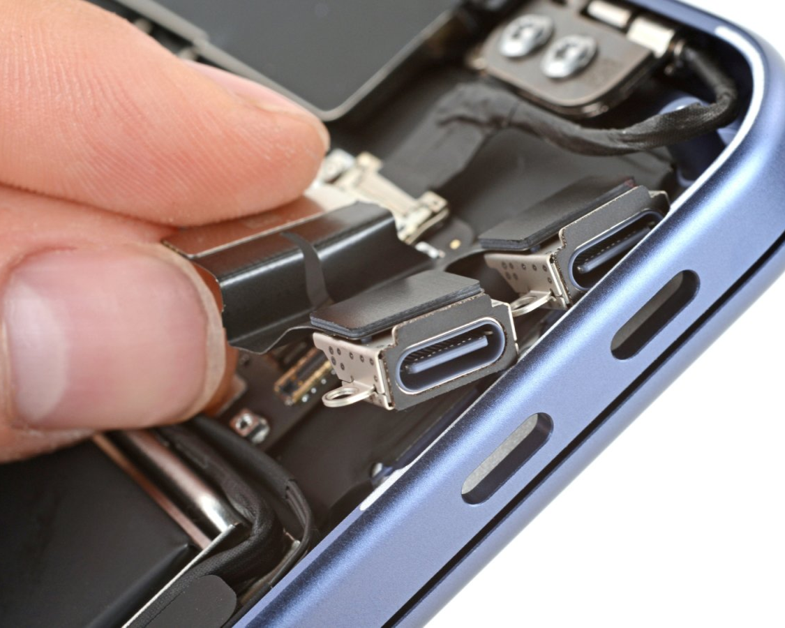 Close-up of a MacBook’s internal USB-C port assembly as a finger lifts the small daughterboard from inside the aluminum frame, showing the modular charging ports and connector cable.