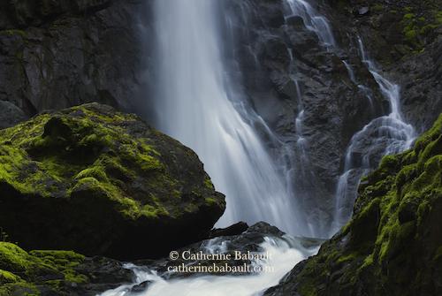 A cascade coming down a rock face and going between moss-covered boulders.