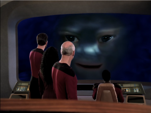 Riker, Troi and Picard stare at a distorted face on the main Enterprise viewscreen.