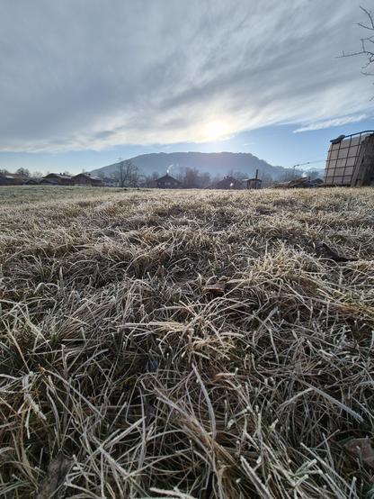 A tranquil and atmospheric rural scene unfolds in this image, capturing the quiet beauty of a frosty morning. The foreground features a field blanketed in a light layer of frost, its grassy surface glistening under the soft light of the rising sun. The frost adds a delicate sparkle, enhancing the peaceful and crisp atmosphere of the scene.

In the background, a cluster of quaint houses nestles at the foot of a rolling hill, their rooftops also touched by frost. The houses, with their rustic cha…