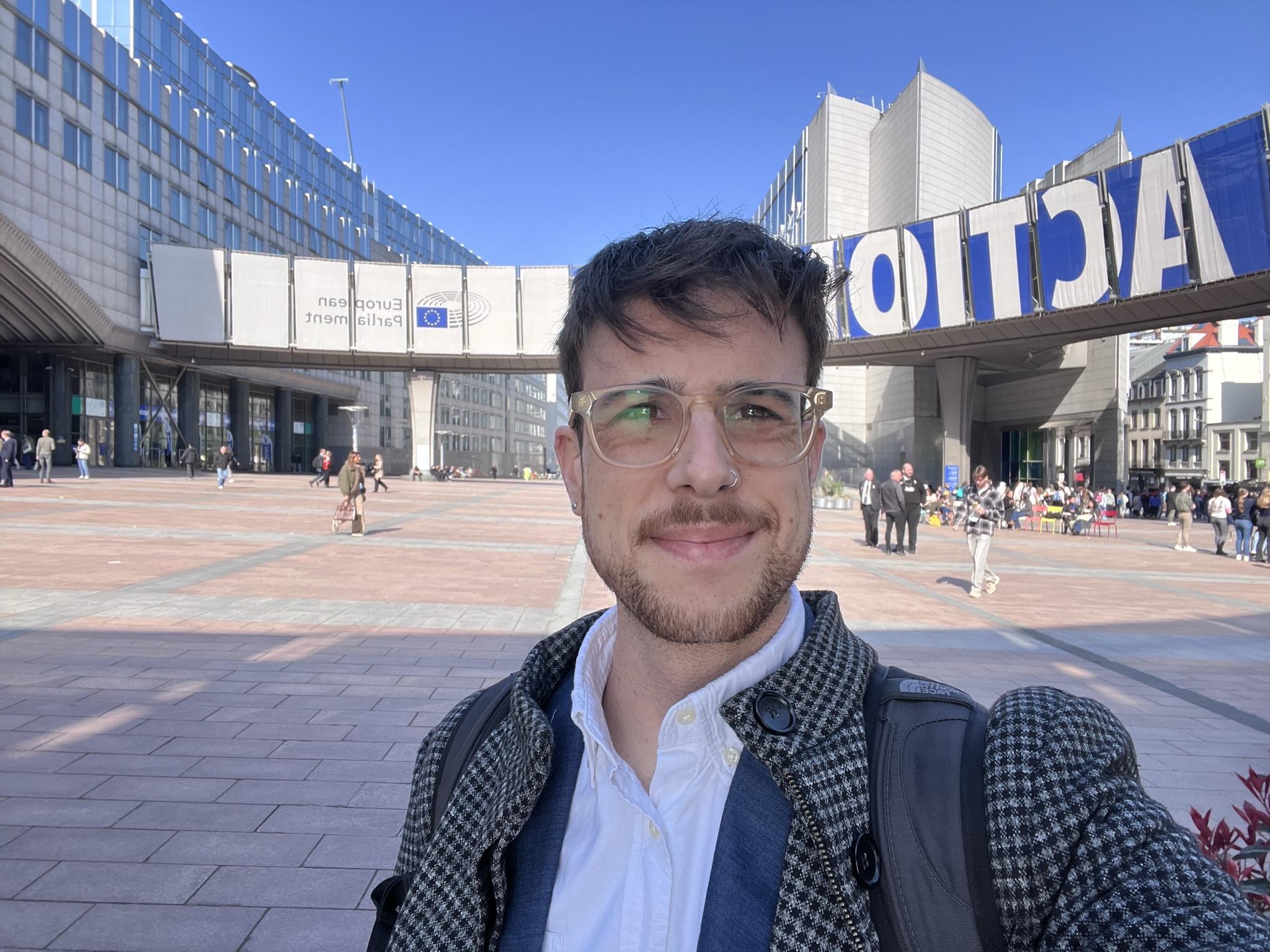 A person stands in the foreground, smiling in front of a large modern building with signage indicating the European Parliament. The area is bustling with people, and the sky is clear and blue.