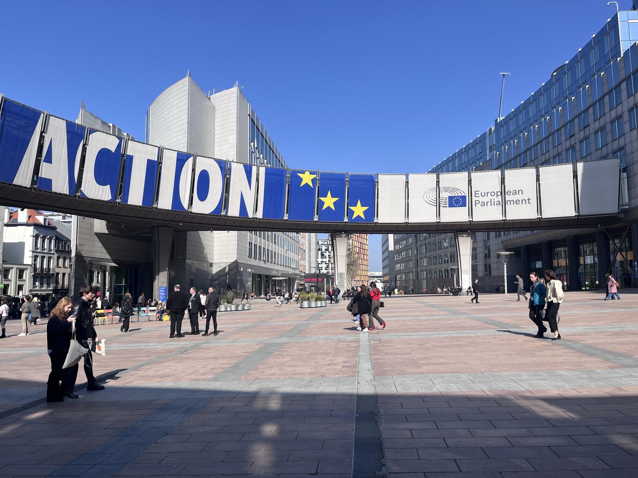 A plaza in front of the European Parliament featuring a large sign that reads "ACTION" and includes European Union flags.