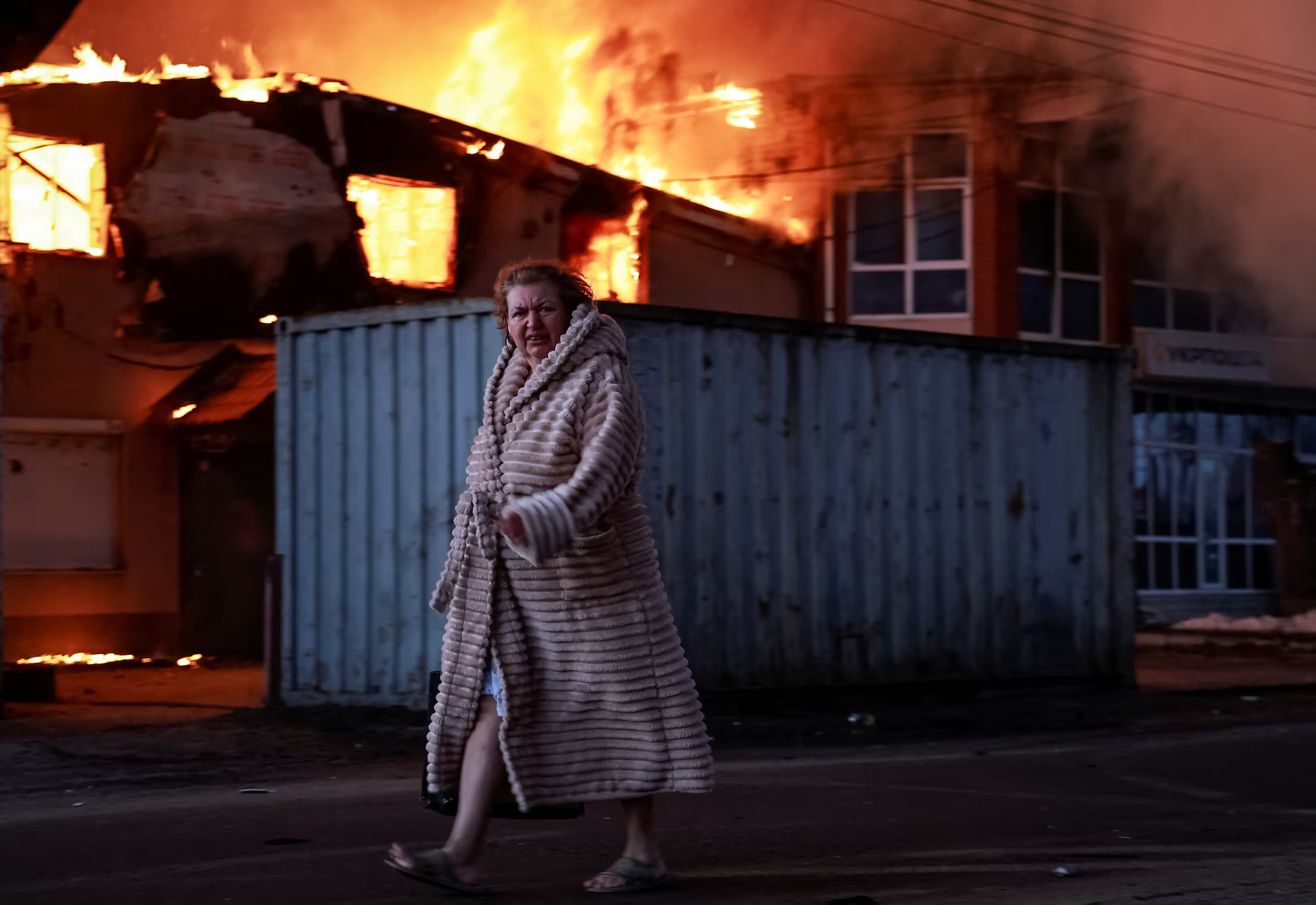 A woman in a gown walks past a burning house.
