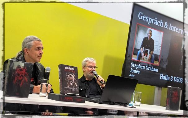 Ein grauhaariger Mann mit Zopf in Lederjacke links, es ist der Autor Stephen Graham Jones, und ein grauhaariger Mann mit Brille, es ist ein Verlagsmitarbeiter auf einer Bühne. Auf dem Tisch vor ihnen steht eine Ausgabe des Buches "The Buffalo Hunter Hunter", das Jones in der Veranstaltung auf der Leipziger Buchmesse vorgestellt hat.