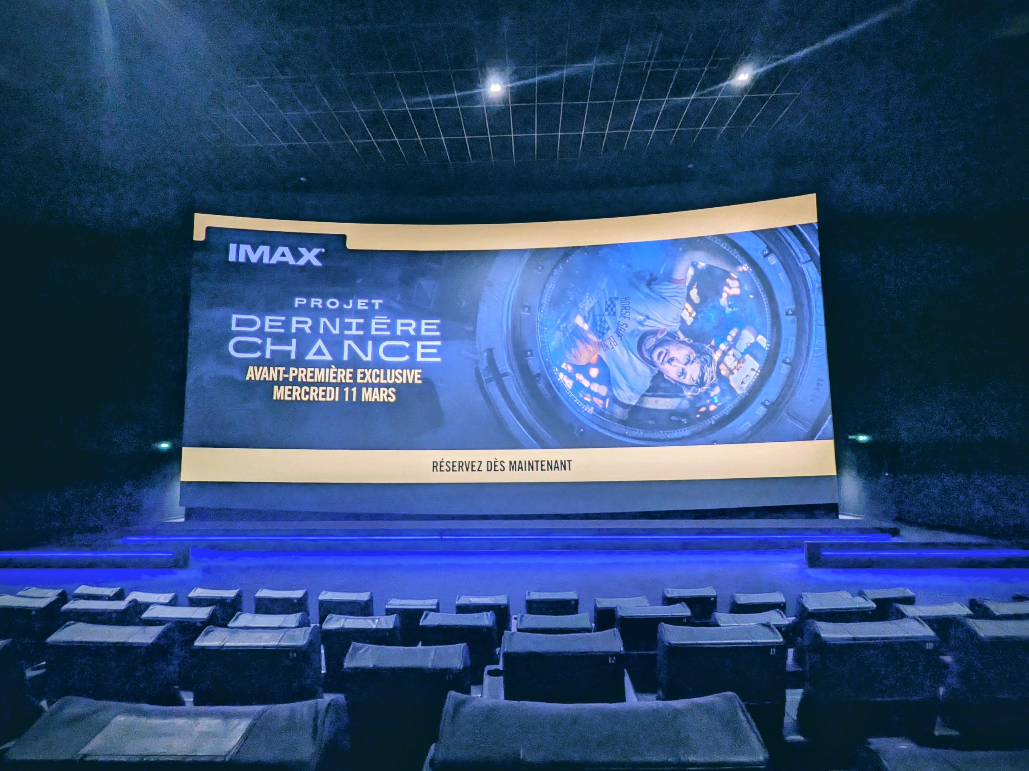 A view from inside a dark movie theater looking toward a large, illuminated IMAX screen. The screen displays an advertisement with the IMAX logo and the text "PROJET DERNIÈRE CHANCE" followed by "AVANT-PREMIÈRE EXCLUSIVE MERCREDI 11 MARS". The bottom edge of the screen has a yellow strip reading "RÉSERVEZ DÈS MAINTENANT". On the right side of the screen, an image shows an astronaut floating inside a circular spacecraft module. In the foreground, rows of empty, dark theater seats face the screen, with a blue light illuminating the walkway at the front.