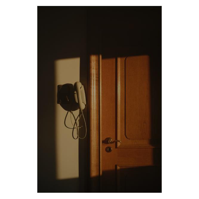 Morgensonne wirft einen rechteckigen Lichtschein auf eine alte Eichentür. Neben der Tür hängt ein altes, graues Haustelefon schief an der Wand. Der Lichtschein wirkt wie ein Rahmen, denn der Rest der Tür und der Wand liegt im tiefen Schatten.