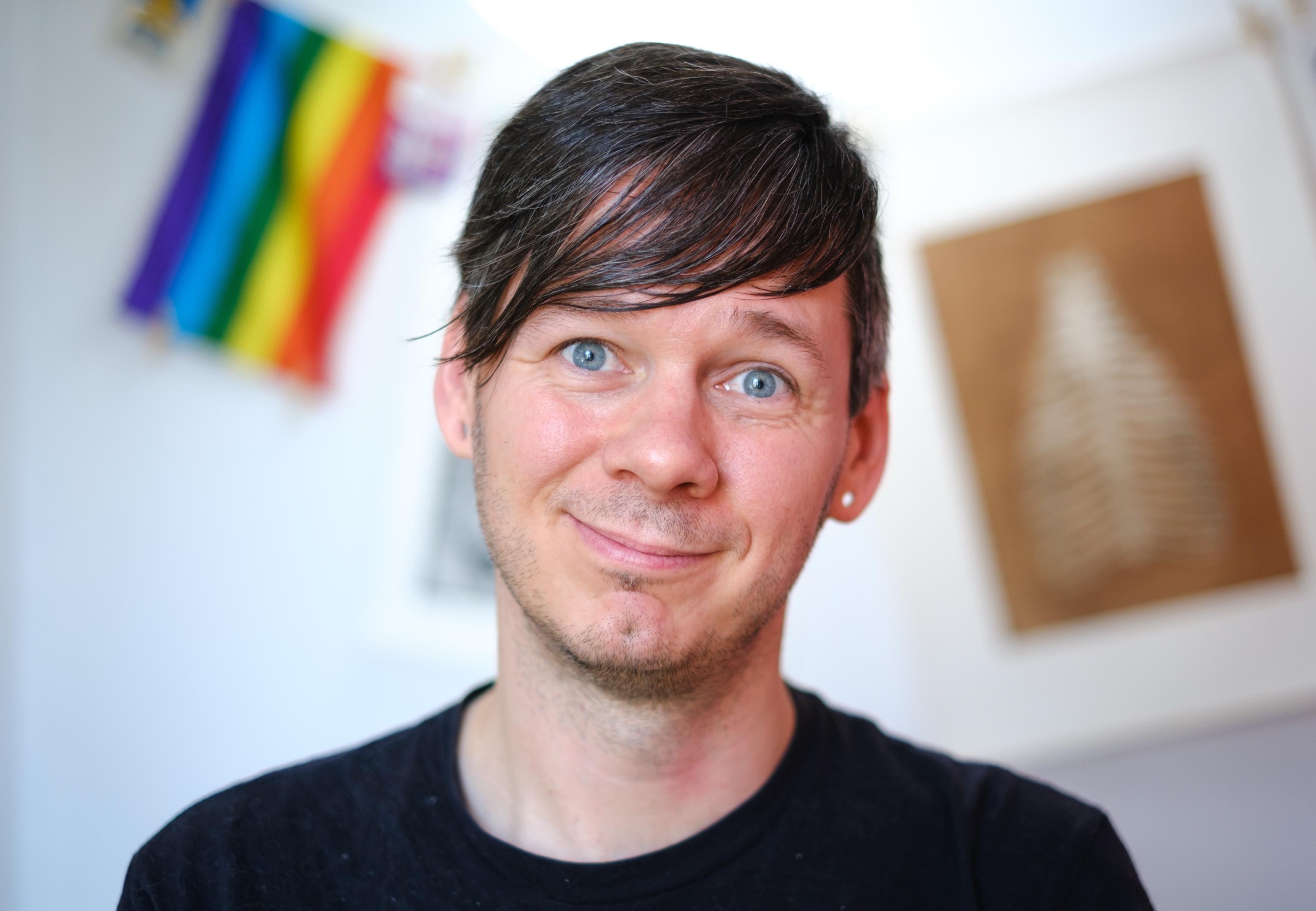 Portrait of a person with blue eyes and dark, emo-style hair. In the background is a blurry pride flag.