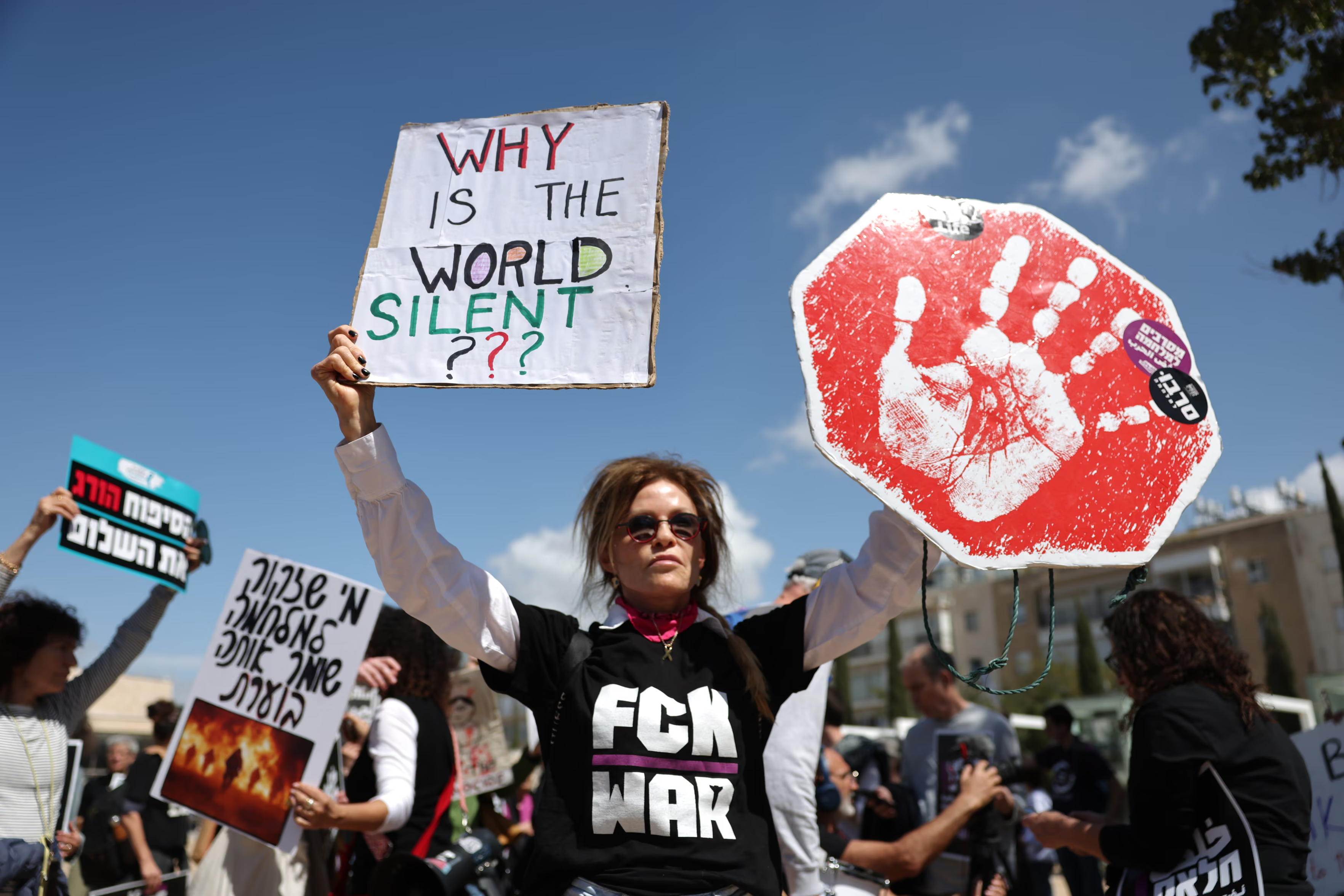 A female Israeli protester in a t-shirt saying FCK WAR is holding a stop sign in one hand and a sign which reads WHY IS THE WORLD SILENT? in the other.