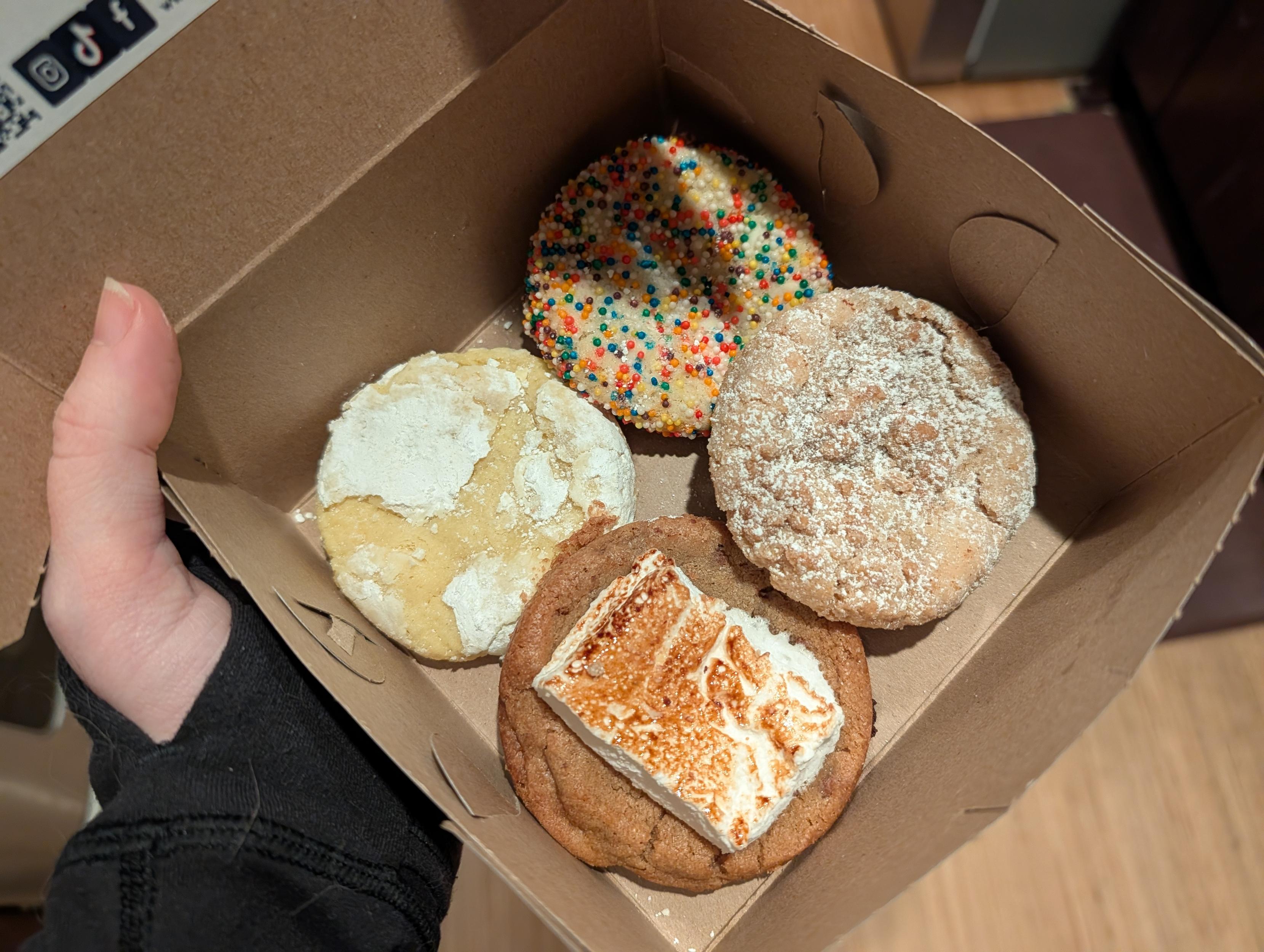 Holding up a box of cookies - there's a lemon crinkle, a smore one, a coffee crumble one, and a birthday cake one. The perfect breakfast