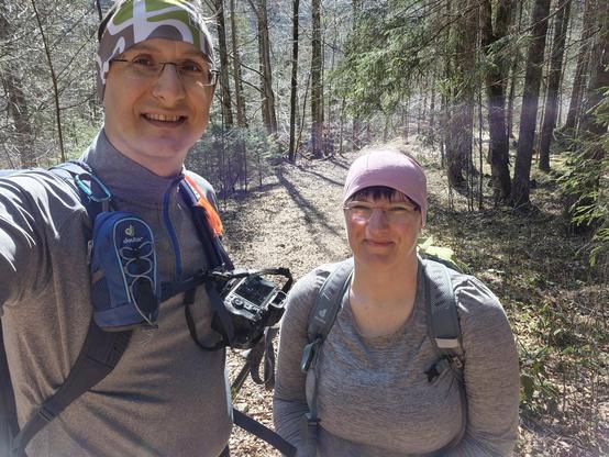 Two hikers smiling into the camera.
The scene is in a woodland.