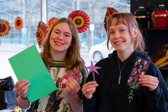 Two younng oman inside a storefront with round paper flowers on the windows,   both have blond hair and hold paper creations that they made