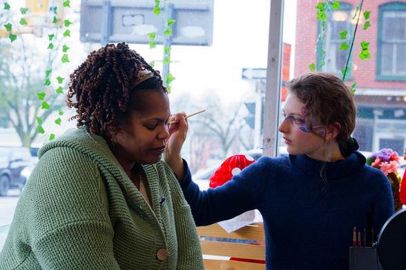 While woman on the left in a blue turtleneck has her hair in a pony tail as she paints the face of a black woman in a grey sweater with tightly curled hair with a glass window behind them showing the street scene outside