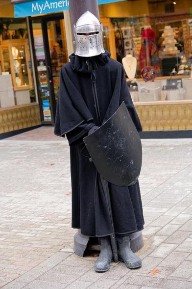 Man with shiny curved metal helmet and black cloak and black shield and dark boots sands straight against light pole in front of gift store