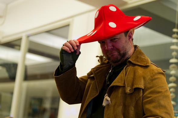 Man in yellow leather coat has a red mushroom cap with white circular spots with a dark indoor storefront with windows and a string of rocks can be seen in the background