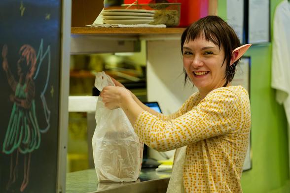 Woman in yellow textured shirt is smiling with elf ears and a short and very straight haircut and i think holding a plastic bag with a box on it with a kitchen on the other side of a window and on the left a fairy with wings has been drawn on a chalkboard and to the left a wall with big framed paperwork on a green wall