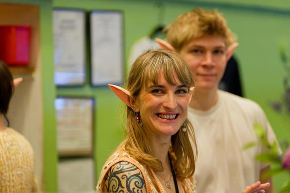 A blonde woman is looking at me and smiling with elf ears and straight and blonde razor cut hair with blurred out young elf man white with a white shirt and short and curly hair behind him with some leaves on the far right and on the left the shoulder and a bit of head withan elf ear pointing out