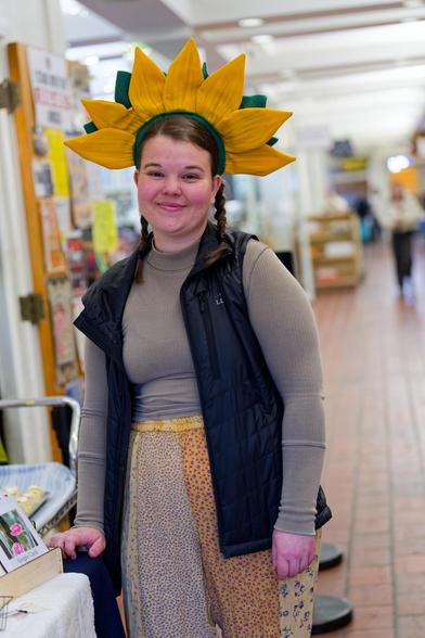 Woman with long hair tied into twin braids with a corona of 7 yellow petals and 5 green leaves is wearing a grey top and dark vest with fine floral pattern pants with a red tile floor and a blurred out store and a distant person inside a store