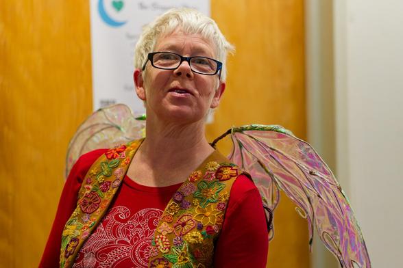 Woman with glasses and short white hair wears insect like wing and a vest with a flower print and red blouse with more white decorations in front of a large door