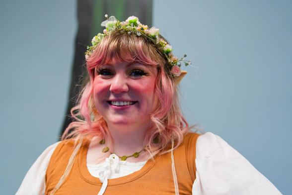 Smiling woman with blonde hair that's been dyed pink in spots near the front is wearing a floral garland and blush makeup and is wearing a white blouse under an orange vest