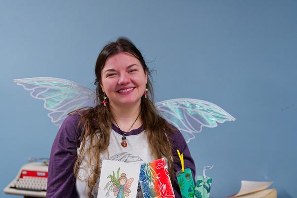 Woman with long wavy hair and a white shirt with purple sleeves is holding books and wearing transparent wings in front of a featureless blue background with an old typewriter in the background